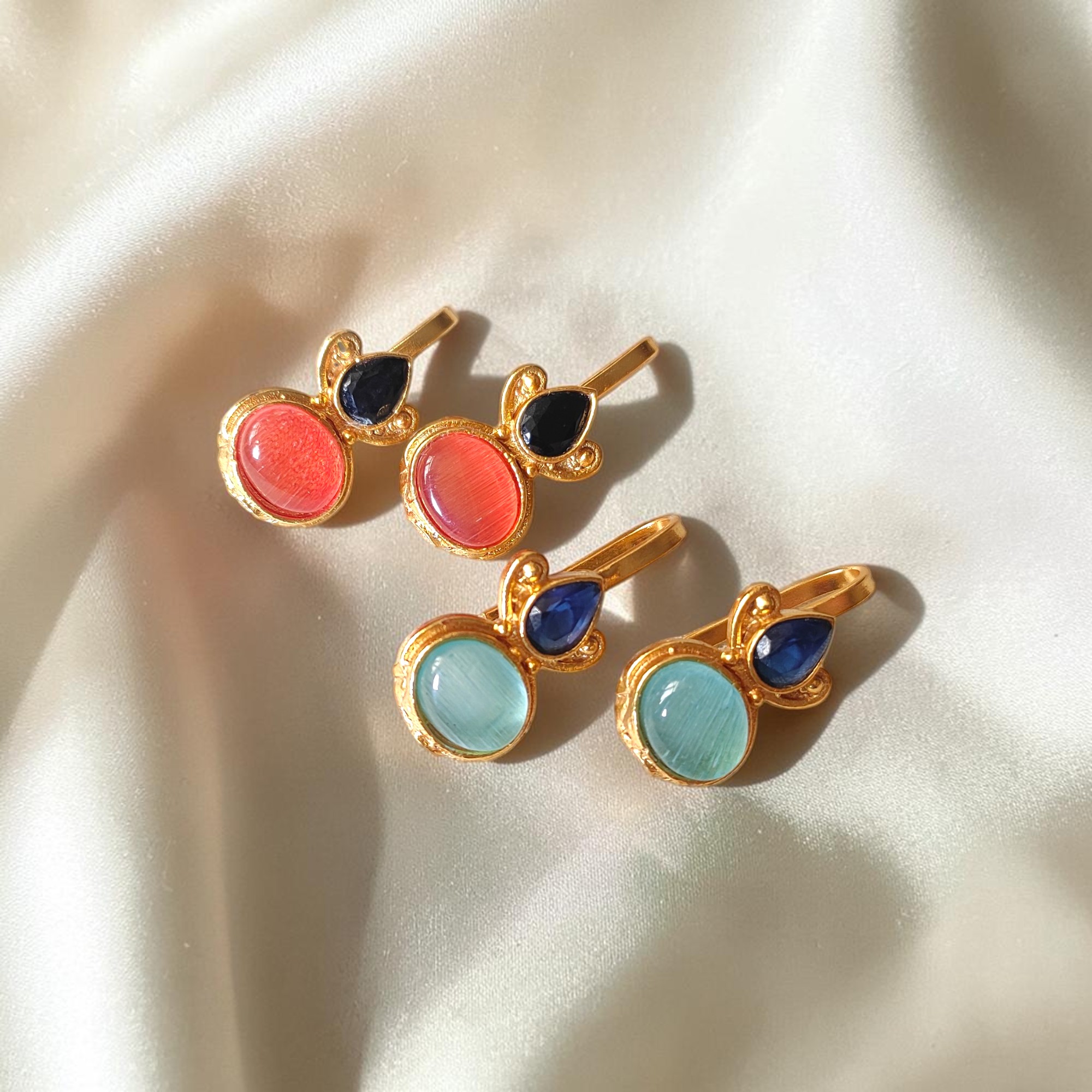 Two pairs of gold plated Bugadi earrings with pink and sky blue Monalisa stones arranged on an ivory satin background.