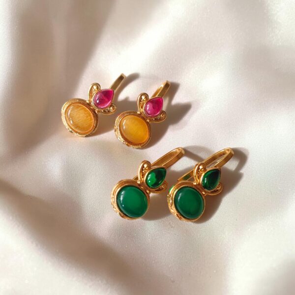 Two pairs of gold-plated Bugadi earrings displayed on ivory satin, with yellow and pink Monalisa stones in one pair and green stones in the other
