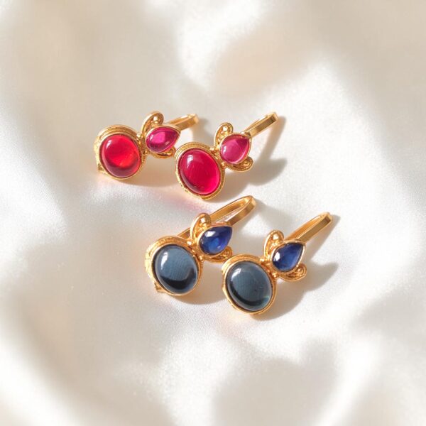 Two pairs of gold plated Bugadi earrings with red and dark blue Monalisa stones arranged on an ivory satin background.