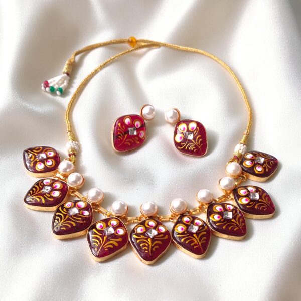 Maroon hand-painted Hasli necklace with floral Tanjore-inspired artwork, pearl detailing, Monalisa stone elements and matching earrings displayed on white satin.