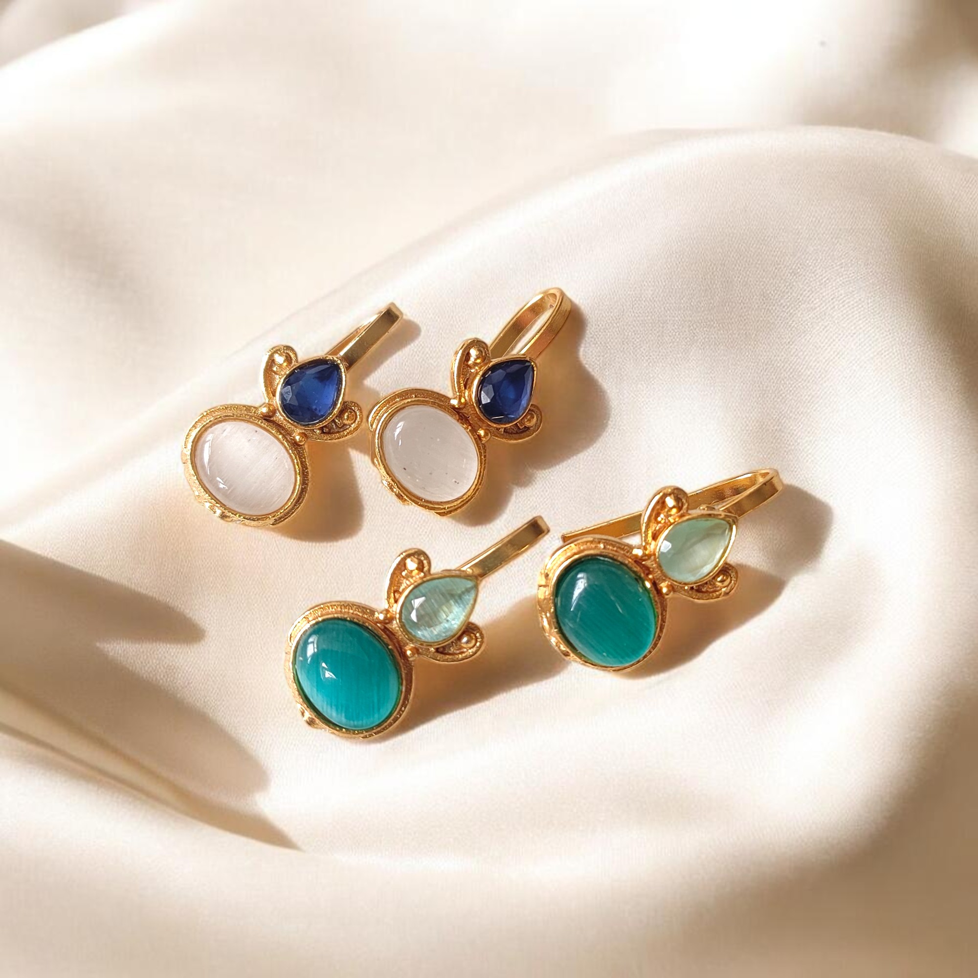 Two pairs of gold plated Bugadi earrings with white and turquoise Monalisa stones arranged on an ivory satin background.